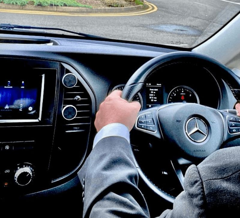 A chauffeur in a dark suit with a hand on the steering wheel of a Mercedes
